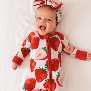 May include: A baby wearing a red and white strawberry print onesie with a red zipper and matching red footies. The baby is wearing a pink and red strawberry print headband. The baby is lying on a white surface and smiling.