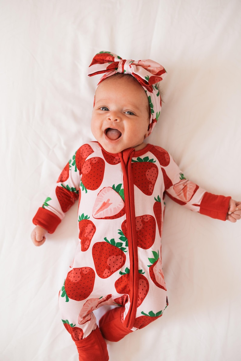 May include: A baby wearing a red and white strawberry print onesie with a red zipper and matching red footies. The baby is wearing a pink and red strawberry print headband. The baby is lying on a white surface and smiling.