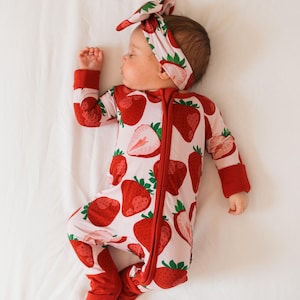 May include: A baby wearing a pink and red strawberry print sleeper with a red bow headband. The sleeper has a zipper down the front and red cuffs on the sleeves and legs.
