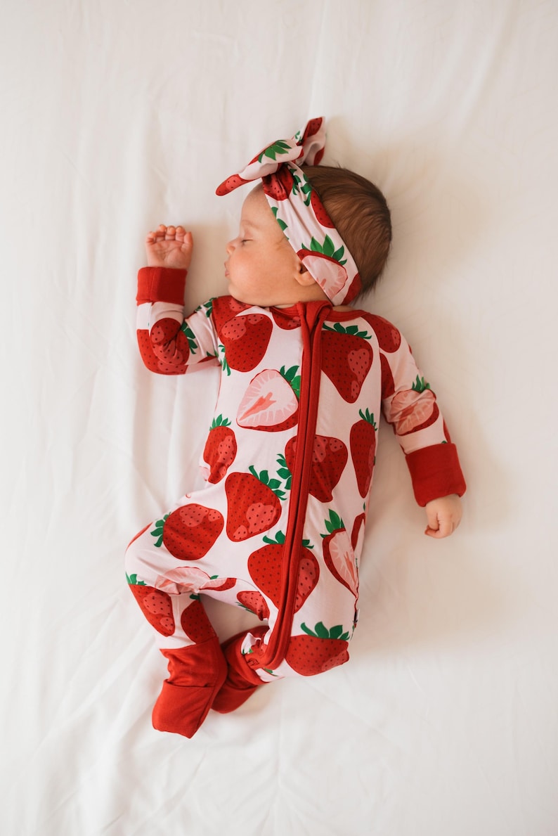 May include: A baby wearing a pink and red strawberry print sleeper with a red bow headband. The sleeper has a zipper down the front and red cuffs on the sleeves and legs.