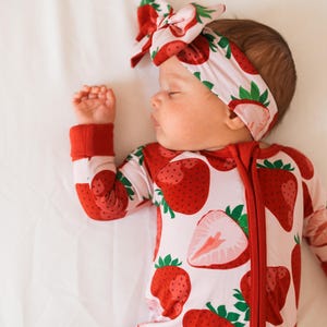May include: A baby wearing a pink and red strawberry print sleeper with a matching headband. The sleeper has a zipper down the front and the headband is tied in a bow.