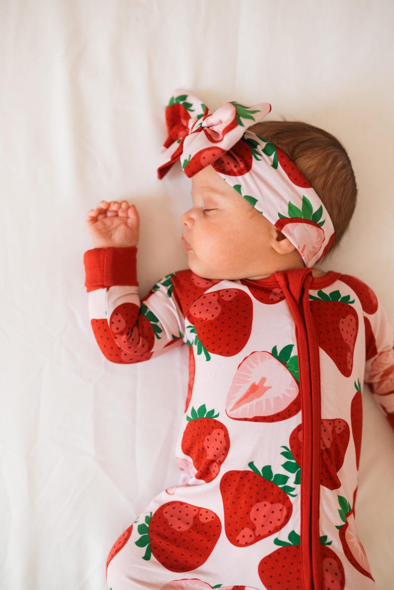 May include: A baby wearing a pink and red strawberry print sleeper with a matching headband. The sleeper has a zipper down the front and the headband is tied in a bow.