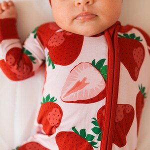 May include: A baby wearing a pink and red strawberry print sleeper with a red zipper. The baby is wearing a white headband with a pink bow.