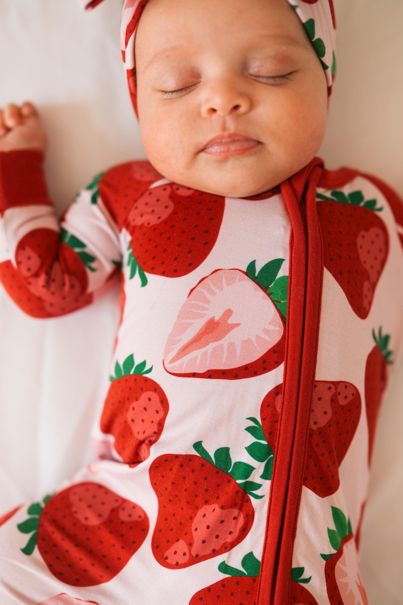 May include: A baby wearing a pink and red strawberry print sleeper with a red zipper. The baby is wearing a white headband with a pink bow.