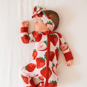 May include: A baby wearing a red and white strawberry print sleeper with a matching headband. The sleeper has a zipper down the front and the headband is tied in a bow.