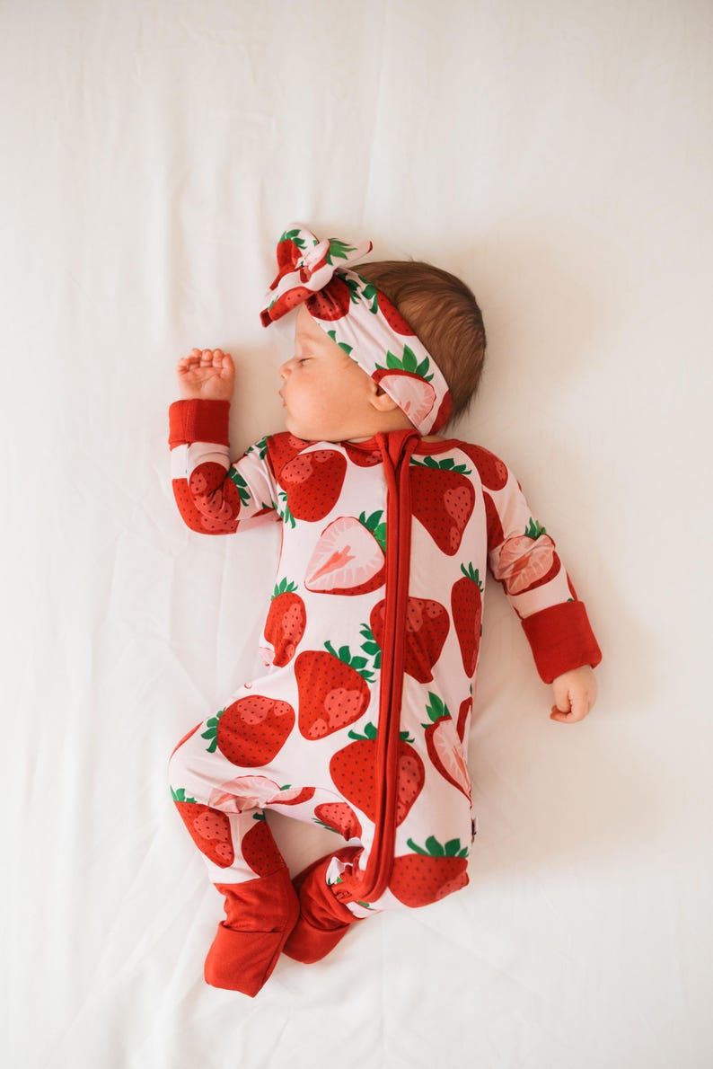 May include: A baby wearing a red and white strawberry print sleeper with a matching headband. The sleeper has a zipper down the front and the headband is tied in a bow.