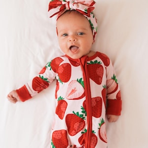May include: A baby wearing a pink and red strawberry print onesie with a matching headband. The onesie has a zipper down the front and the headband is tied in a bow.