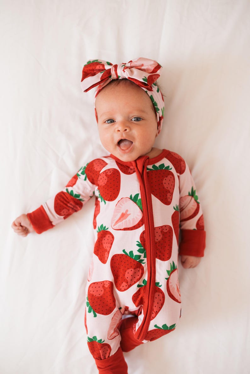 May include: A baby wearing a pink and red strawberry print onesie with a matching headband. The onesie has a zipper down the front and the headband is tied in a bow.