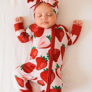May include: A baby wearing a pink and red strawberry print sleeper with a matching strawberry print headband. The sleeper has a zipper down the front and the headband is tied in a bow.
