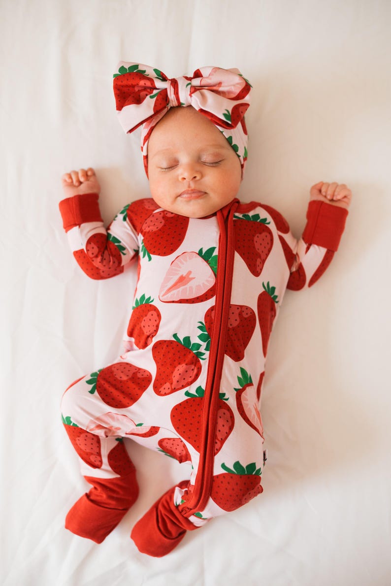 May include: A baby wearing a pink and red strawberry print sleeper with a matching strawberry print headband. The sleeper has a zipper down the front and the headband is tied in a bow.