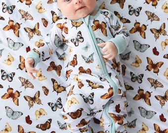 May include: A baby in a white onesie and headband, patterned with butterflies. The onesie has a zip and pale blue trim. The baby lies on a white blanket, also decorated with butterfly illustrations.
