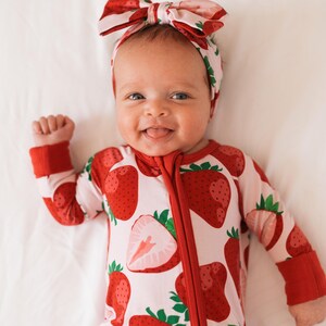 May include: A baby wearing a red and white strawberry print sleeper with a red zipper and red cuffs. The baby is wearing a pink and red strawberry print headband with a bow.
