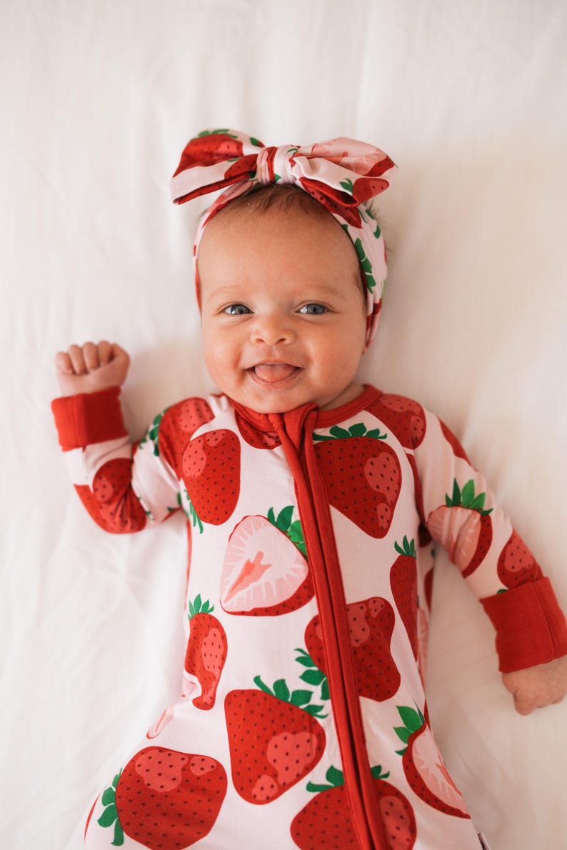 May include: A baby wearing a red and white strawberry print sleeper with a red zipper and red cuffs. The baby is wearing a pink and red strawberry print headband with a bow.