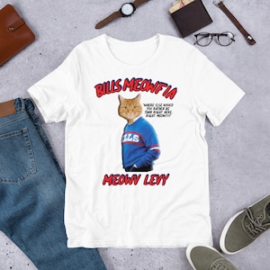 May include: White t-shirt with a graphic of a cat wearing a blue and red football jersey. The text "BILLS MEOWFIA" is above the cat and "MEOWV LEVY" is below the cat. The text "WHERE ELSE WOULD YOU RATHER BE THAN RIGHT HERE. RIGHT MEOW!!!" is to the right of the cat.