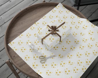 Puede incluir: Un regalo envuelto con una cinta y un lazo marrón se encuentra sobre papel de regalo blanco con un patrón de estrellas doradas. Unas tijeras doradas y un objeto de vidrio transparente también están sobre el papel, sobre una mesa de madera.