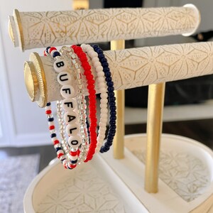 May include: A set of five beaded bracelets on a gold stand. The bracelets are red, white, and blue, and one has the word "BUFFALO" spelled out in black beads.