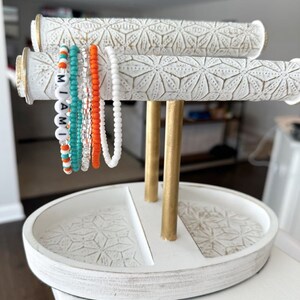 May include: White ceramic jewelry stand with two gold metal posts and a round tray. The stand has a decorative pattern of white stars on a white background. The tray is divided into two sections. Five colorful beaded bracelets are displayed on the stand. The bracelets spell out the word "MIAMI" in white beads.