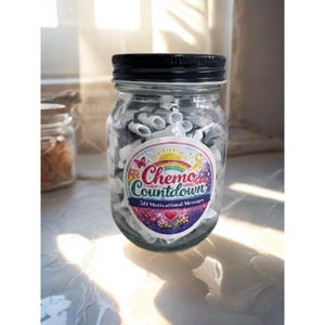Op de afbeelding: Een heldere glazen pot met een zwarte deksel, gevuld met opgerold wit papier. Een kleurrijk etiket op de pot leest "Chemo Countdown" met "50 Motivational Messages". De pot staat op een reflecterend oppervlak.