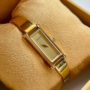 Puede incluir: Un reloj Gucci dorado con una esfera rectangular y una correa dorada. La esfera del reloj es de color beige claro con la palabra "GUCCI" impresa. El reloj se exhibe sobre un cojín de color dorado.