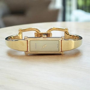Puede incluir: Un reloj Gucci dorado con una esfera rectangular y una correa dorada. La esfera del reloj es de color crema claro con agujas doradas y el logotipo de Gucci. El reloj se muestra sobre una superficie de madera de color claro.