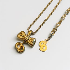 May include: Gold-coloured necklace with a bow pendant and 'CD' logo. The chain is fine, and the necklace has a lobster clasp. The bow pendant is the main feature, adding a touch of elegance to the jewellery.
