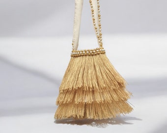 Puede incluir: Un pequeño bolso de borlas beige con una correa de tela blanca y cuentas doradas y blancas. El bolso tiene un diseño de borlas en capas.