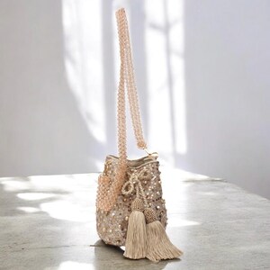 Handmade Wayuu crossbody bucket bag in beige, decorated with shimmering sequins, pearls, and tassels. The long beaded strap allows it to be worn comfortably on the shoulder or crossbody, while the artisan crochet base ensures durability and unique character. A versatile beaded bag perfect for special occasions, gifting, or adding a touch of luxury to everyday outfits.