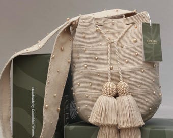 Puede incluir: Un bolso tipo cubo de ganchillo beige con cuentas perladas dispersas, una correa larga para el hombro y un cierre de cordón con dos borlas grandes. Una etiqueta rectangular verde con el texto FOLHA está unida al lado del bolso.