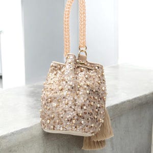 Wayuu handmade sequin bucket bag with long beaded strap hanging naturally, showing its crossbody functionality. Beige crochet base decorated with pearls, sequins, and tassels highlights artisan techniques. Perfectly versatile as a crossbody, shoulder, or occasion bag. A slow fashion piece crafted in Colombia with attention to detail and timeless elegance.