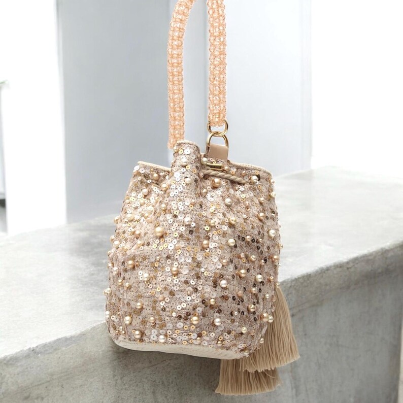 Wayuu handmade sequin bucket bag with long beaded strap hanging naturally, showing its crossbody functionality. Beige crochet base decorated with pearls, sequins, and tassels highlights artisan techniques. Perfectly versatile as a crossbody, shoulder, or occasion bag. A slow fashion piece crafted in Colombia with attention to detail and timeless elegance.