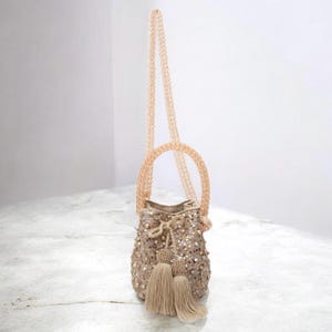 Medium handmade Wayuu bucket bag resting on a stone surface, with long beaded strap extending upward. The beige crochet bag is hand-decorated with sequins, pearls, and tassels, reflecting artisan craftsmanship. A delicate crossbody or shoulder bag option, perfect for weddings, anniversaries, and gifting. Luxury slow fashion accessory from Colombia.