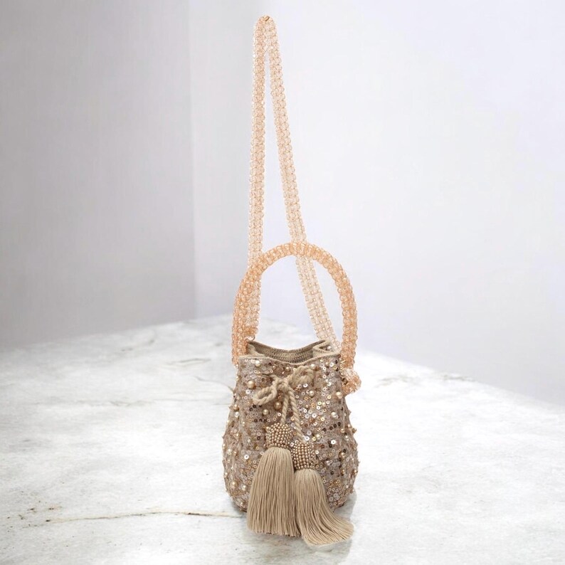 Medium handmade Wayuu bucket bag resting on a stone surface, with long beaded strap extending upward. The beige crochet bag is hand-decorated with sequins, pearls, and tassels, reflecting artisan craftsmanship. A delicate crossbody or shoulder bag option, perfect for weddings, anniversaries, and gifting. Luxury slow fashion accessory from Colombia.