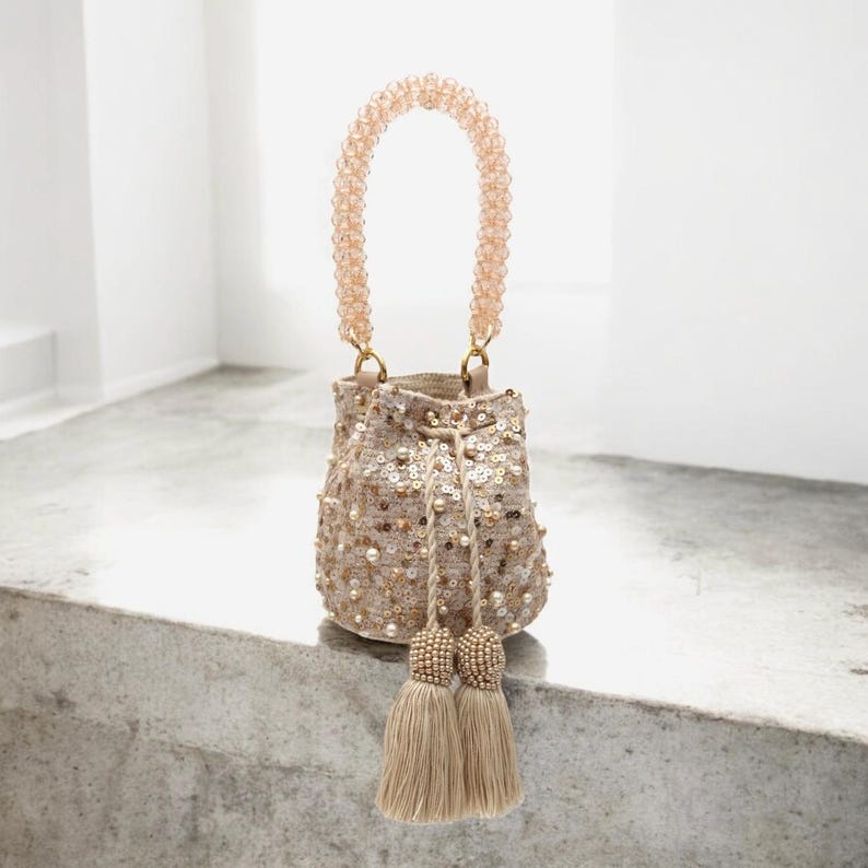 Front view of handmade Wayuu beaded bucket bag with short strap, designed for use as a chic handheld purse. The beige crochet base is richly decorated with sequins, pearls, and stones, paired with tassels for an elegant finish. A one-of-a-kind artisan handbag, blending traditional Wayuu weaving with modern luxury details, perfect for weddings or evening style.