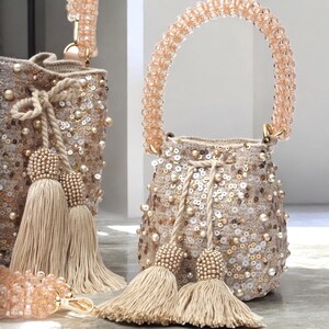 Close-up of two artisan Wayuu sequin bucket bags, each with a short beaded strap for handheld carrying. The beige crochet base is fully hand-embellished with sequins, pearls, and stones, finished with oversized tassels. Shiny and elegant, these handmade bags highlight their double strap design for versatile use. Perfect luxury handbags for weddings, parties, or boho chic fashion.