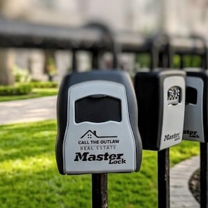May include: A row of black and silver Master Lock key lock boxes hanging on a dark metal fence. Each box features a combination dial, a protective cover, and the printed text Call The Outlaw Real Estate above the Master Lock brand name logo.