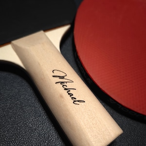 May include: Close-up of a ping pong paddle with a wooden handle. The handle is light-colored with the name "Michael" printed in black script. The paddle's red rubber surface is visible in the background, along with a black surface.