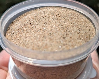 May include: A small clear plastic container with a white lid filled with a light brown powder.