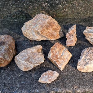May include: A collection of various-sized, rough-textured rocks in shades of tan and brown. The stones are arranged on a dark, textured surface, with sunlight casting shadows. The rocks vary in shape and size, showcasing natural geological formations.