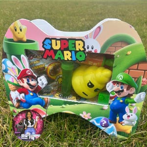 May include: A Super Mario-themed gift box with a controller shape, featuring Mario and Luigi characters. The box includes a yellow character, green tubes, and the text "Super Mario". The background shows a green grassy area.