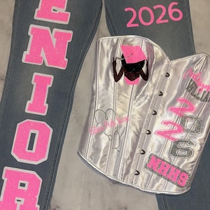 Op de afbeelding: Lichtblauwe spijkerbroek met het woord SENIOR in grote roze glitterletters op het been en CLASS OF 2026 in het roze. Een wit satijnen korset-top met een roze afstudeerhoed-afbeelding, de tekst Future Pediatric Nurse en de letters NHS.