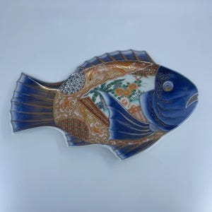 Puede incluir: Un plato decorativo de cerámica con forma de pez. El plato presenta una cabeza y cola azules, con intrincados patrones naranjas, dorados y blancos en el cuerpo. El diseño incluye motivos florales y geométricos.