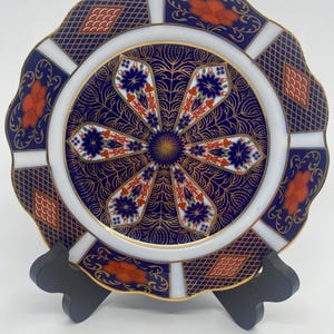 May include: A decorative plate with a scalloped edge, featuring a navy blue background with intricate gold filigree and floral patterns. The plate has white and orange accents, and a gold rim. It is displayed on a black stand.
