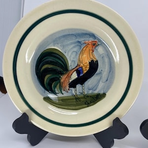 May include: A ceramic plate featuring a hand-painted rooster design. The rooster has orange, black, and blue feathers, with a green and blue background. The plate has a cream-colored base with a green border.