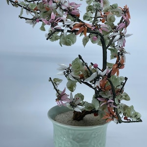 May include: A decorative bonsai tree with a light green ceramic pot. The tree features a dark brown trunk and branches adorned with pink, white, and orange flowers and green leaves. The pot has a detailed floral design.