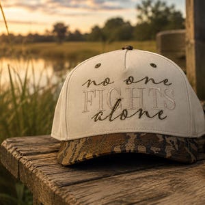 May include: A cream-colored baseball cap with a camouflage-patterned brim. The front of the cap features the embroidered text 'no one FIGHTS alone' in a combination of cursive and block lettering. The cap has a structured crown and eyelets.
