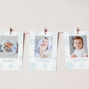 Op de afbeelding: Drie witte fotokaarten met blauwe sneeuwvlokontwerpen en foto's van een baby op verschillende leeftijden. De kaarten zijn gelabeld met "Nieuwgeboren", "6 maanden" en "1 jaar oud!"