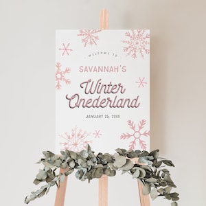 Op de afbeelding: Een welkomstbord op een houten ezel met de tekst "Welcome to Savannah's Winter Onederland" in roze en zwart. Roze sneeuwvlokken versieren de witte achtergrond. Eucalyptustakken zijn aan de basis van het bord gedrapeerd.