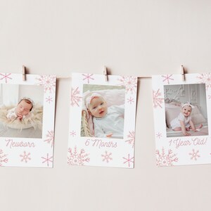 Op de afbeelding: Drie witte fotokaarten met roze sneeuwvlokdesigns en een babyfoto op elke kaart. De kaarten zijn opgehangen met touw en wasknijpers. De tekst op de kaarten luidt "Pasgeboren", "6 maanden" en "1 jaar oud!"