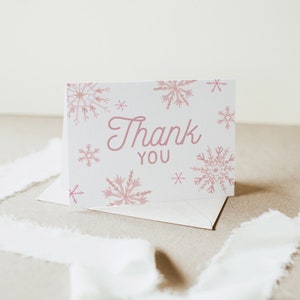 Op de afbeelding: Een witte kaart met een roze sneeuwvlokkenpatroon en de tekst "Thank You" in roze cursief schrift. De kaart ligt op een witte envelop.