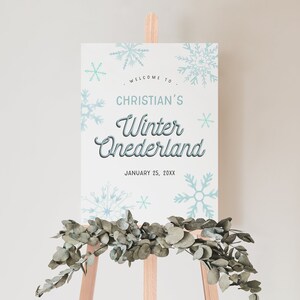 Op de afbeelding: Een wit bord met blauwe sneeuwvlokken en de tekst "Welkom bij Christian's Winter Wonderland, 25 januari 20XX" op een ezel met groen.